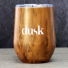 A wooden-textured tumbler with dusk written in white on the front, sitting on a smooth surface against a dark background.