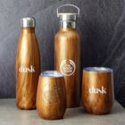 Four insulated bottles and cups with a wood grain design are displayed against a gray textured background. Two are labeled dusk and two THE BODY SHOP. They vary in size and shape, including bottles with lids and stemless cups.