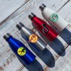 Four stainless steel water bottles lie on a wooden surface. They are blue, silver, red, and white, each featuring different logos: Lacoste, Hard Rock Café, TikTok, and Starbucks.