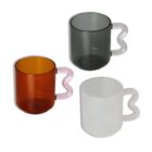 Three translucent mugs with heart-shaped handles are displayed. The top mug is gray with a white handle, the left mug is amber with a pink handle, and the bottom mug is white with a white handle. The mugs have a modern design.