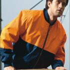 A person wearing an orange and navy work jacket is holding onto a wooden object. The word WORK WEAR is printed at the bottom, and ABCINI is at the top. The background is blurred.