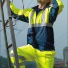 A person wearing a bright yellow and navy work outfit stands at the base of a ladder, adjusting a yellow hard hat. The person is looking upwards, with a cityscape in the background. The text on the image reads TBOCINI WORK WEAR.