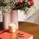 A lit candle in a small holder sits on a wooden table beside a red book with a gold design. A tall vase with red flowers and white babys breath is in the background.
