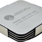 A stack of square coasters with rounded corners, held together by a metal holder. The top coaster has the text Hydraulic and Pneumatic Pty Ltd with contact information printed on it. The coasters have a sleek, modern design.