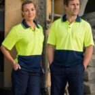 Two people wearing yellow and navy blue work uniforms stand in front of a large, muddy construction vehicle. They both appear confident and are standing with hands in pockets.
