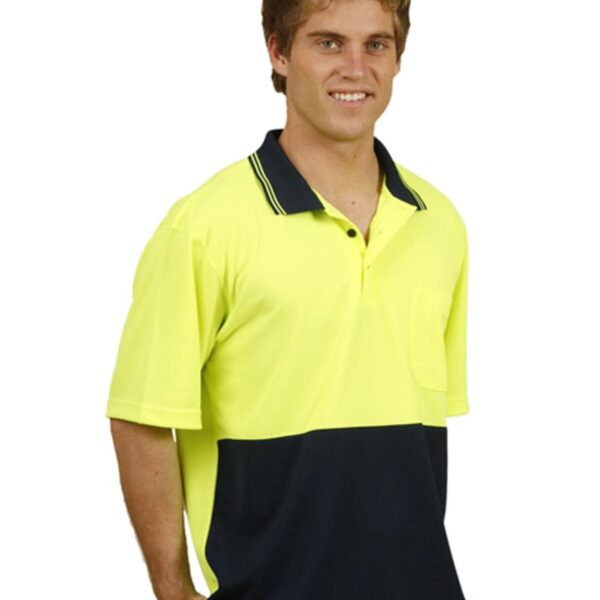 A person wearing a bright yellow and dark blue polo shirt with a pocket, standing and smiling. The shirt has a collar with dark stripes. The sleeves are short, and they are wearing light blue jeans.