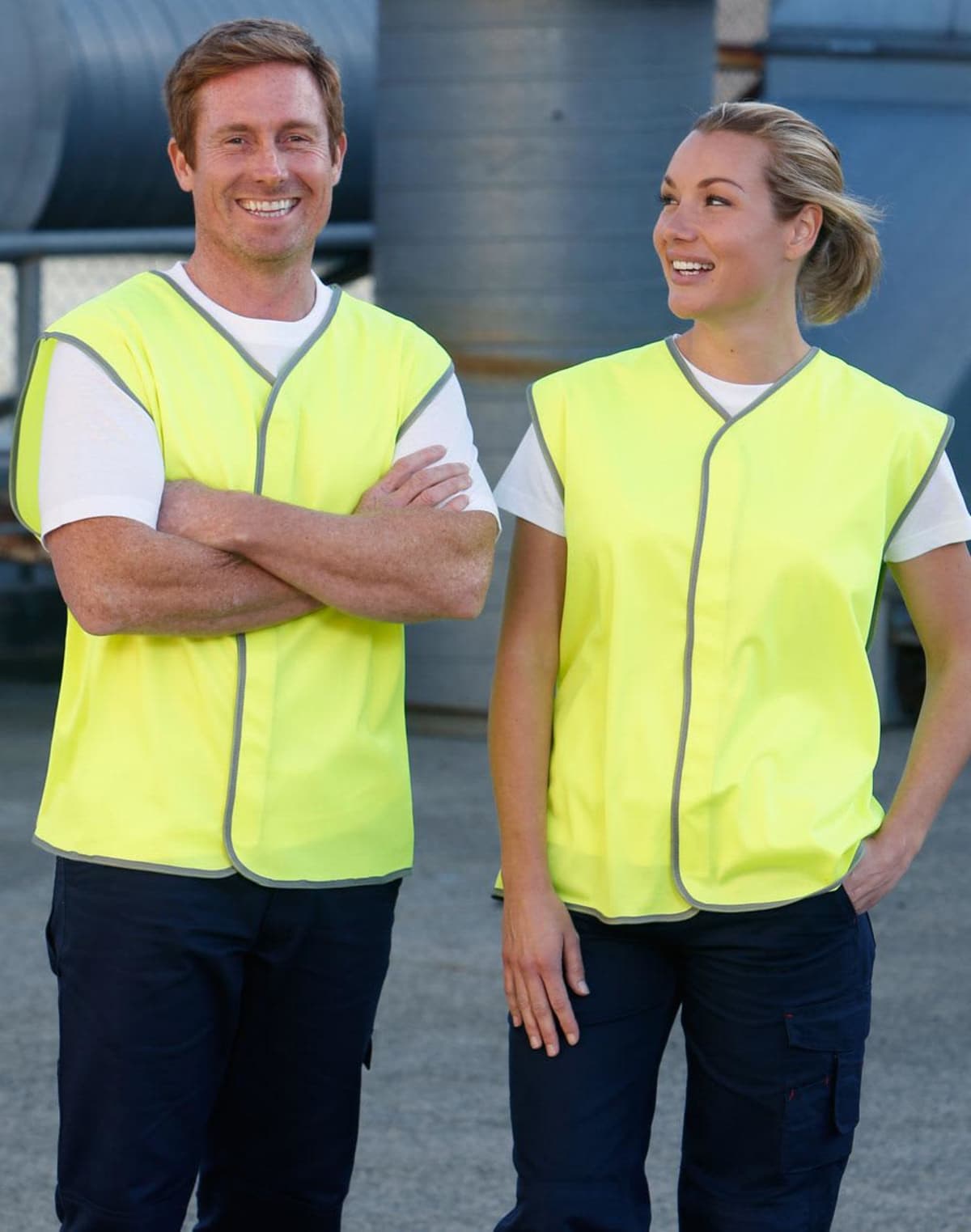 Two people in fluorescent yellow safety vests stand outdoors. The man has his arms crossed and is smiling, while the woman looks at him, also smiling. They appear to be near industrial equipment.