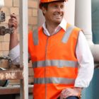 A man wearing an orange reflective safety vest and hard hat stands smiling near industrial equipment. He is holding a lever with one hand and has a relaxed posture, indicating a work environment. The background features pipes and a brick wall.