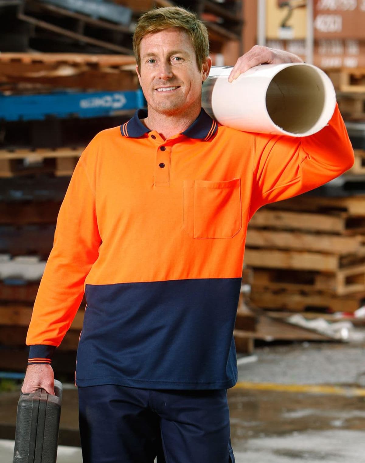 A man in a bright orange and navy work shirt stands confidently. He holds a large white pipe on his shoulder and a toolbox in his other hand. Wooden pallets are stacked in the background.