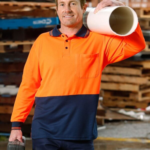 A person in a bright orange and navy blue work shirt holds a white pipe on one shoulder and a toolbox in the other hand. They are standing outdoors with wooden pallets in the background.