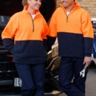 Two people in matching orange and navy uniforms stand by a car with an open hood. Both are smiling and leaning against the vehicle. One person holds a wrench. They are outdoors, with a brick wall in the background.