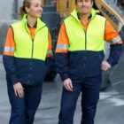 Two construction workers wearing bright yellow high-visibility jackets stand together. The background features a blurred construction vehicle and a concrete surface.