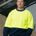A man wearing safety glasses and a high-visibility yellow and navy-blue sweatshirt stands in an industrial setting. He has short brown hair and is smiling slightly. A metal structure and a pile of sand are visible in the background.