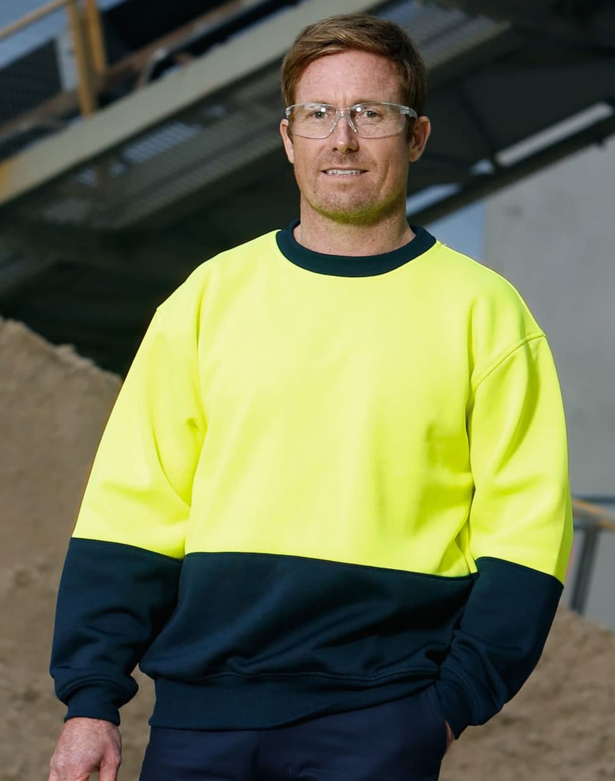 A man wearing safety glasses and a high-visibility yellow and navy-blue sweatshirt stands in an industrial setting. He has short brown hair and is smiling slightly. A metal structure and a pile of sand are visible in the background.