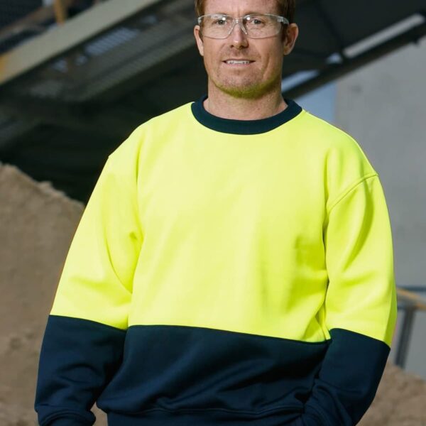 A person wearing safety glasses and a high-visibility yellow and black sweatshirt is standing outdoors in an industrial setting. They have short hair and appear to be in front of a metal structure and a pile of dirt or sand.