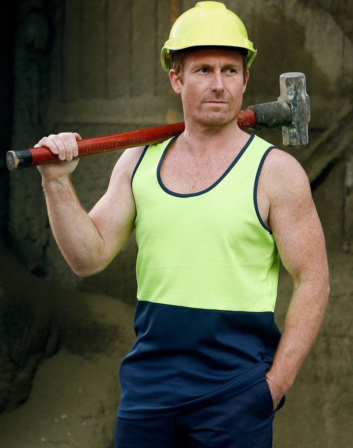 A construction worker in a yellow hard hat and a neon green and navy tank top stands holding a sledgehammer over his shoulder, looking into the distance, with one hand in his pocket. The background is a construction site.