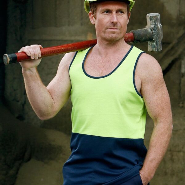 A man wearing a yellow hard hat and a sleeveless neon green and navy shirt stands confidently, holding a sledgehammer over his shoulder. The background is an industrial setting with concrete surroundings.