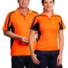 Two people wearing bright orange and black polo shirts stand side by side. The person on the left is smiling and has short hair, while the person on the right has long hair tied back. Both are wearing dark pants.