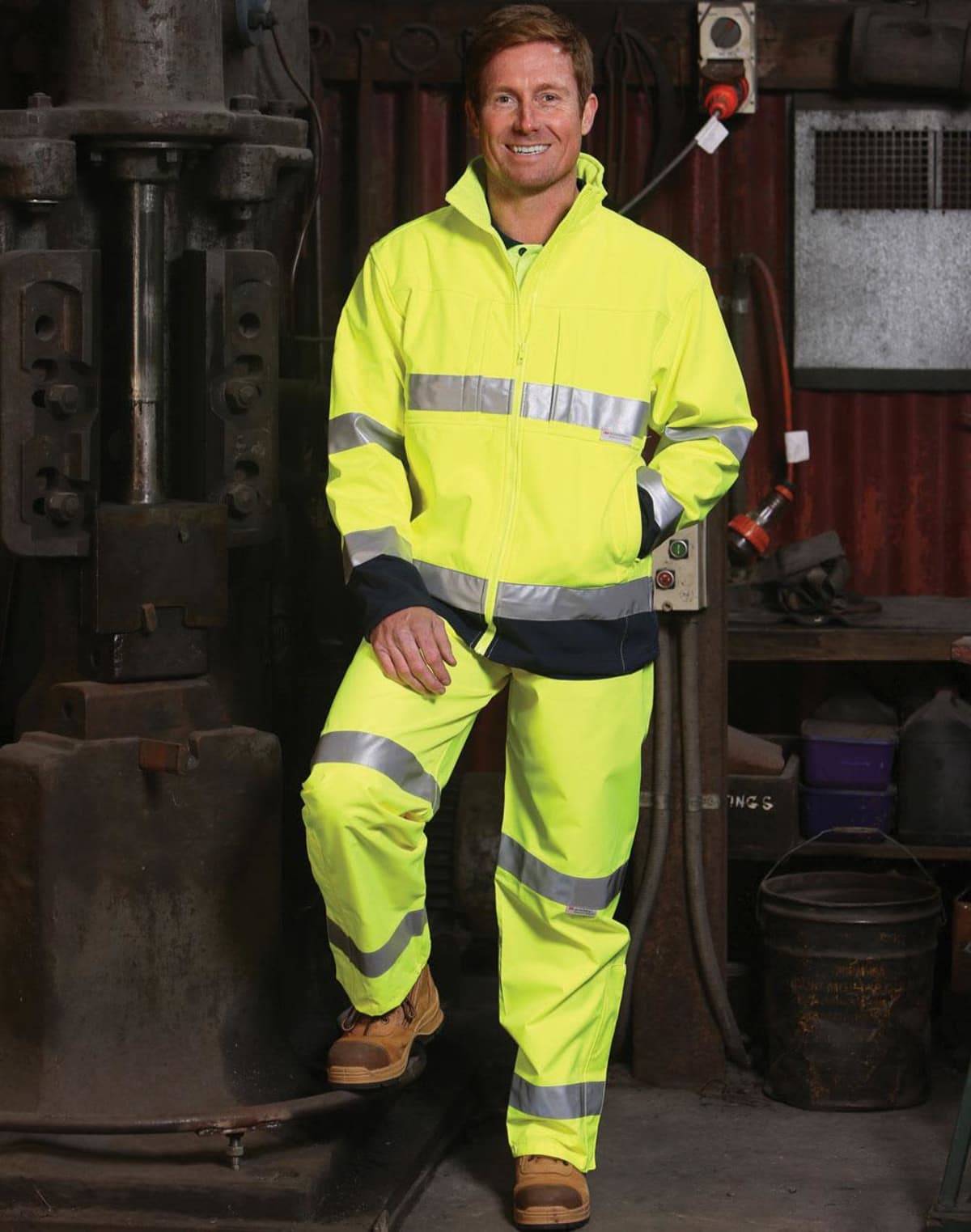 A person wearing a bright yellow safety suit with reflective strips stands in an industrial setting, smiling. They are leaning against machinery, with tools and equipment visible in the background.