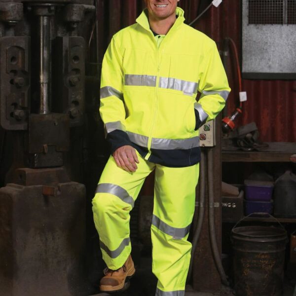 A person wearing a yellow high-visibility safety suit stands in an industrial setting. They are smiling and have one foot resting on a black barrel. The background includes machinery and equipment.