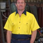 A person with short red hair is wearing a yellow and navy work shirt, standing in a workshop. The background includes industrial equipment and tools.