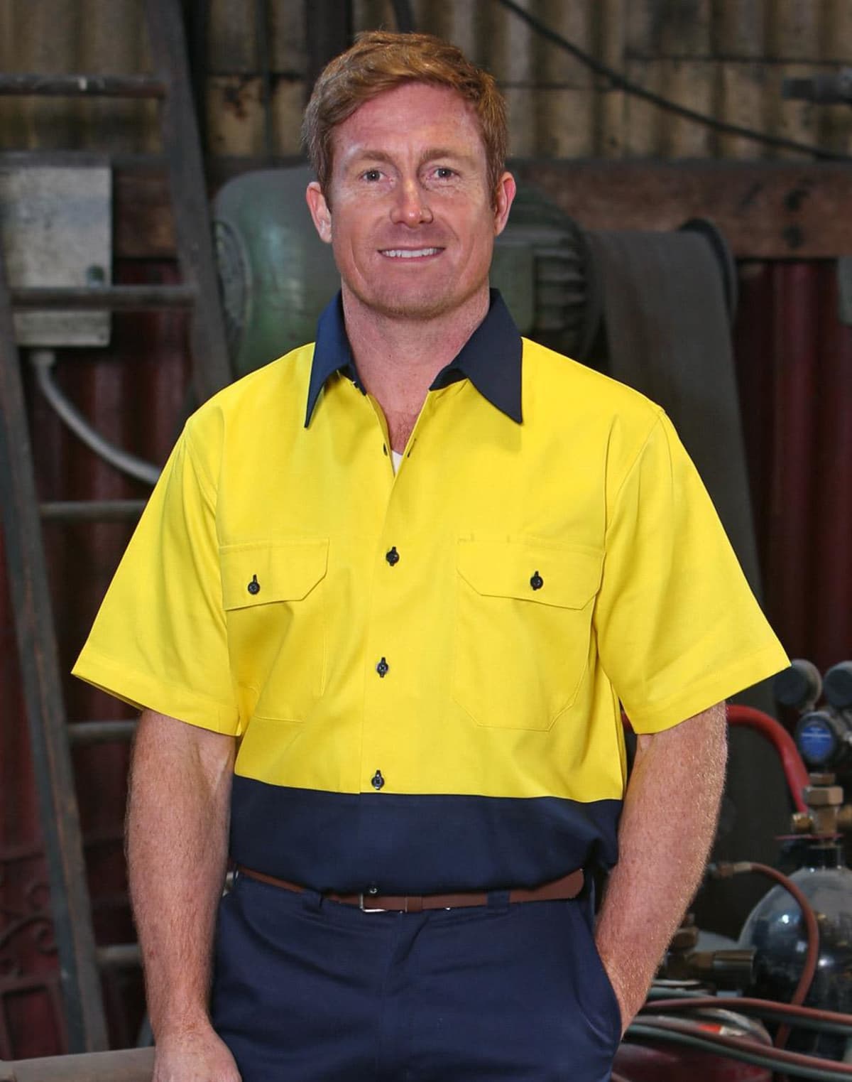 A person with short red hair is wearing a yellow and navy work shirt, standing in a workshop. The background includes industrial equipment and tools.