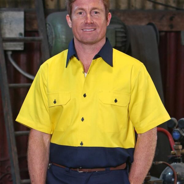A person wearing a yellow and navy work shirt stands in a workshop with equipment in the background.