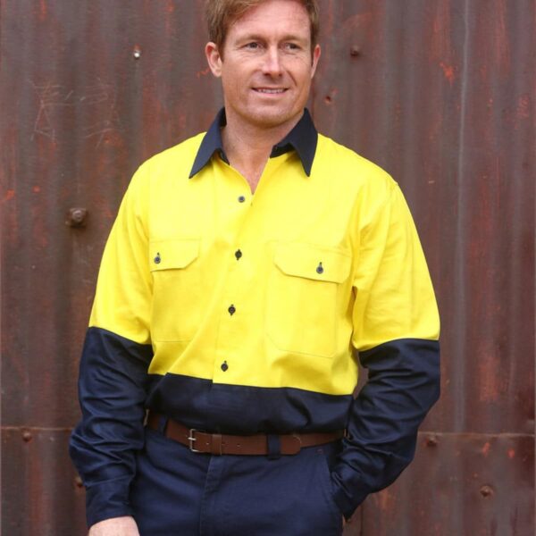 A person wearing a yellow and navy blue work uniform stands in front of a rusty metal wall. They have short hair and are smiling slightly, with hands in their pockets.