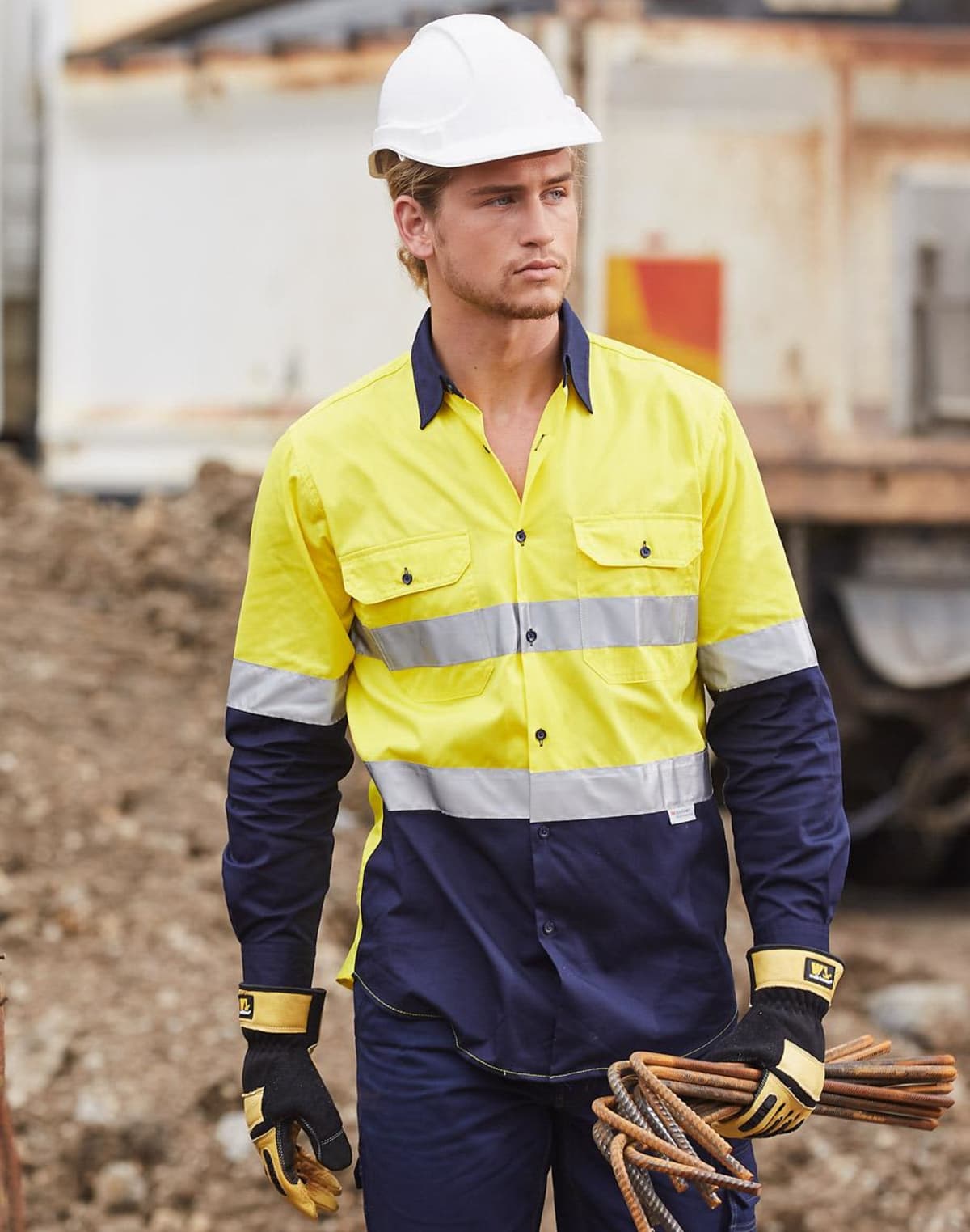 A construction worker wearing a white helmet, a yellow and navy reflective shirt, and gloves is holding metal rods at a construction site. Dirt and construction materials are visible in the background.
