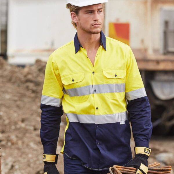 A construction worker in a yellow and navy high-visibility shirt and white hard hat is standing outdoors holding metal rods. He is wearing protective gloves, and there is construction equipment and dirt in the background.