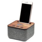 A smartphone is docking into a square speaker with a wooden top and fabric-covered sides. The speaker has three buttons on the front, and a slot for the phone. The phones screen is off. The speaker has a retro rainbow logo.