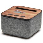 A compact, square speaker with a wooden top panel and grey fabric covering. It has a slot on top and three buttons on the front: power, play/pause, and volume. The top features colorful text.