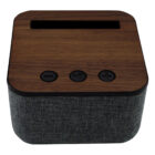 A small, square speaker with a wooden top and three circular buttons: volume down, power, and volume up. The sides are covered in dark gray fabric.