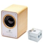Wooden Bluetooth speaker with a cylindrical black speaker in the center of a white front panel. Accompanied by its packaging box on the right, showing product information and illustrations. Modern and minimalist design.