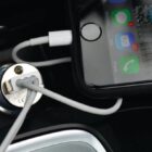Close-up of a smartphone with a black case connected to a car charger via a white cable. The phones screen shows various app icons and notifications. The charger is plugged into a car power outlet.