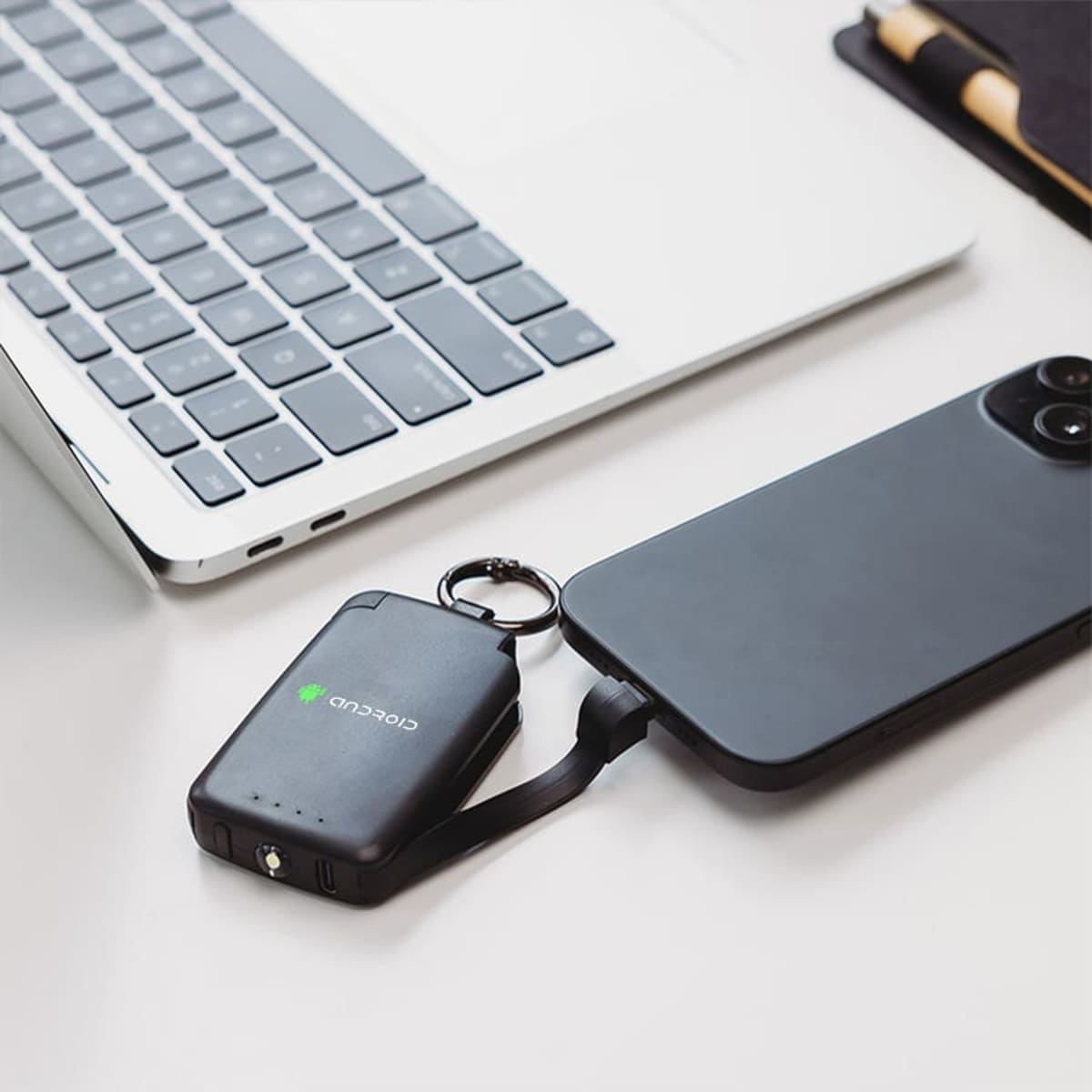 A laptop is on a white desk, with a smartphone connected to a compact black keychain power bank via a short cable. The power bank displays a green logo. Nearby, a notebook is partially visible.