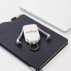 A multi-connector keychain with the Royal Canin logo sits on a closed dark notebook. A corner of a silver laptop is visible in the background, all placed on a white surface.