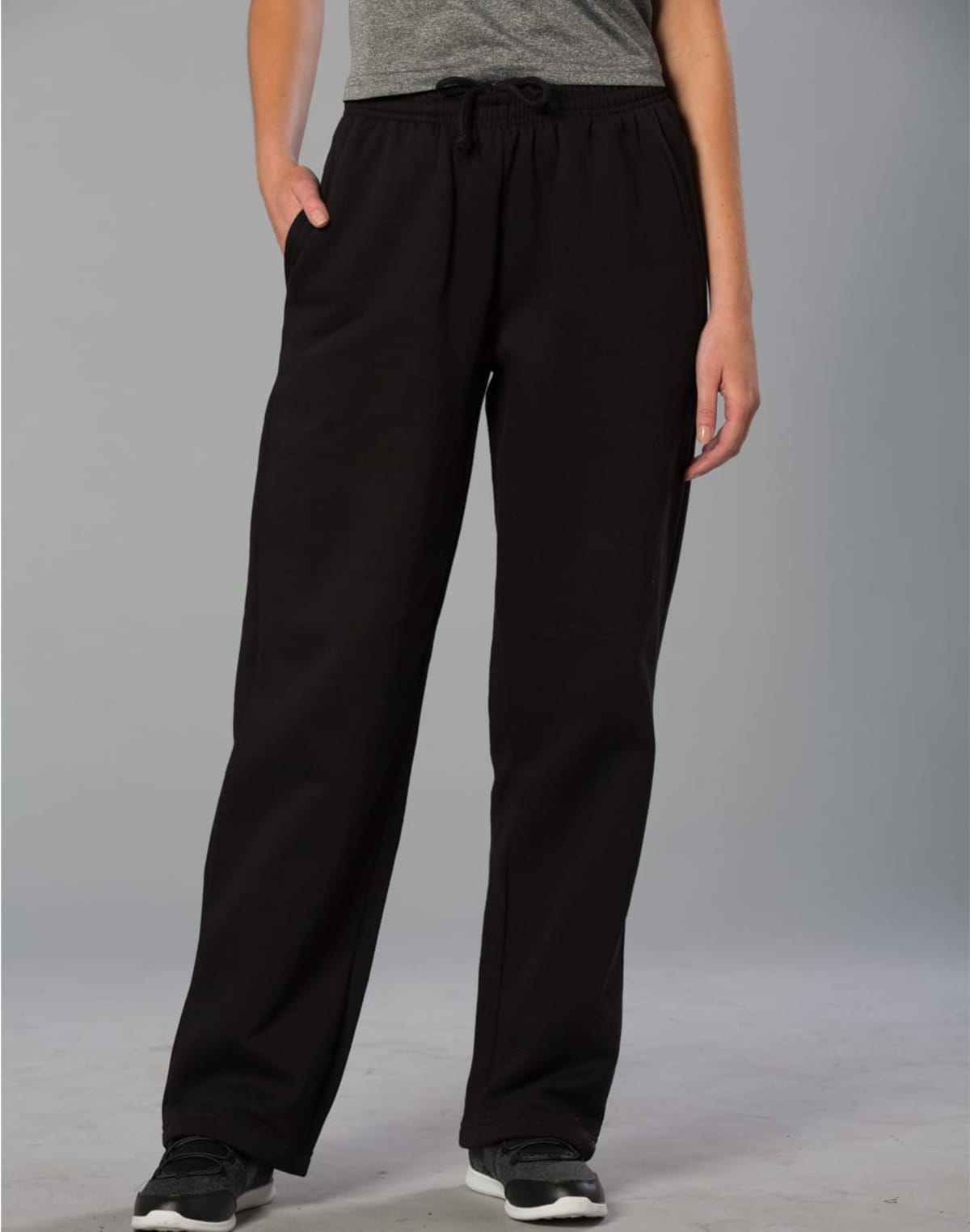 A person wearing black sweatpants and a gray top stands with hands in pockets. The pants have a relaxed fit with a drawstring waist, and the person is wearing black shoes. The background is a plain gray.