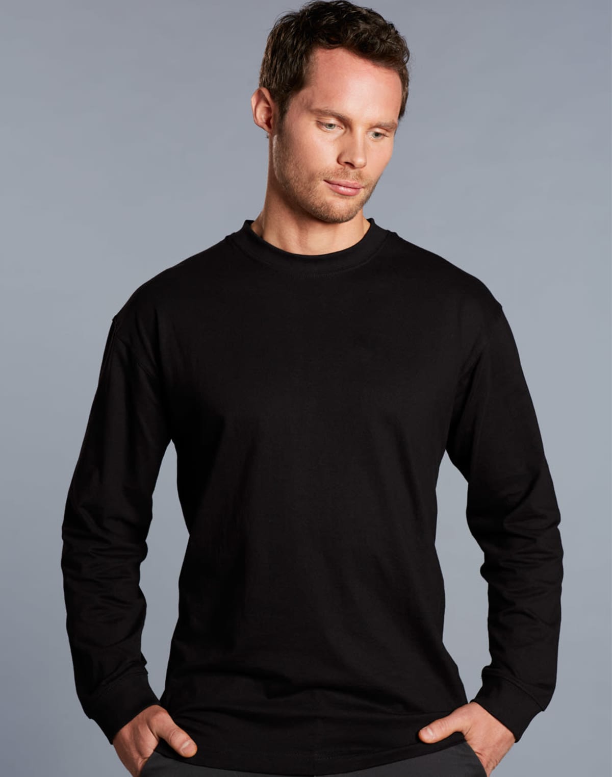 A person with short brown hair wearing a black long-sleeve shirt stands against a plain gray background, looking slightly downwards with a neutral expression.