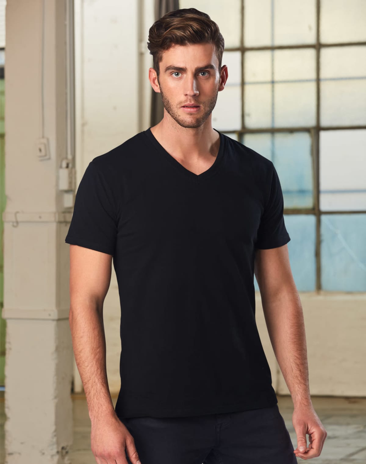 A man with short dark hair is standing indoors wearing a plain black V-neck T-shirt. The background features large windows and industrial-style decor.
