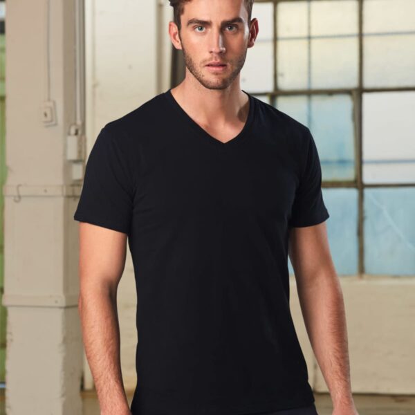 A man with short hair wearing a black V-neck T-shirt stands indoors. The background features large industrial-style windows and white pillars, creating a minimalistic setting.