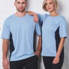 A man and woman stand against a white brick wall wearing matching light blue and black athletic shirts. The woman has short blonde hair, and the man has short dark hair. Both are looking forward with neutral expressions.