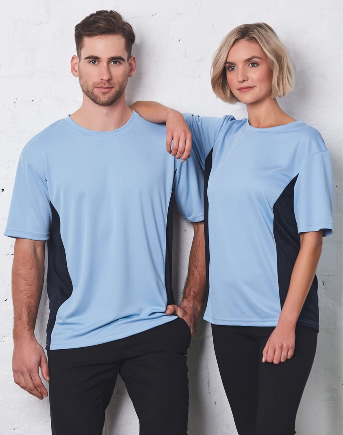 A man and woman stand against a white brick wall wearing matching light blue and black athletic shirts. The woman has short blonde hair, and the man has short dark hair. Both are looking forward with neutral expressions.