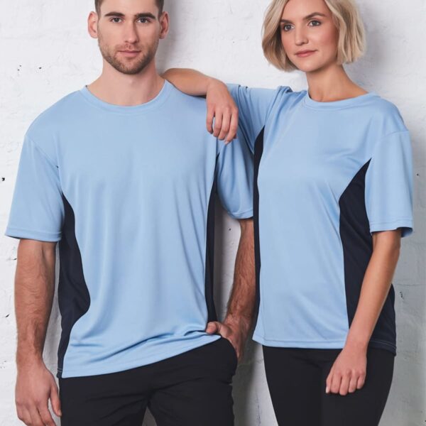 A man and a woman stand side by side against a white brick wall, both wearing light blue T-shirts with black side panels and black pants. The woman rests her arm on the mans shoulder.