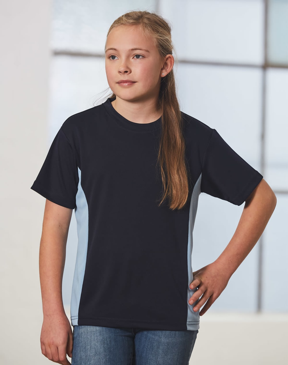A young girl with long blonde hair stands confidently with one hand on her hip, wearing a navy and light blue t-shirt and jeans. She is indoors with a blurred window in the background.