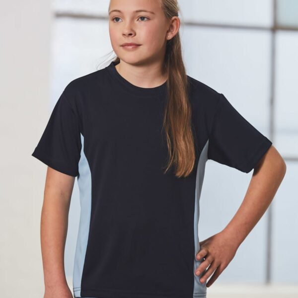 A young girl with long hair stands indoors by a window, wearing a black T-shirt with light blue side panels and blue jeans. She has one hand on her hip and is looking slightly to the side.