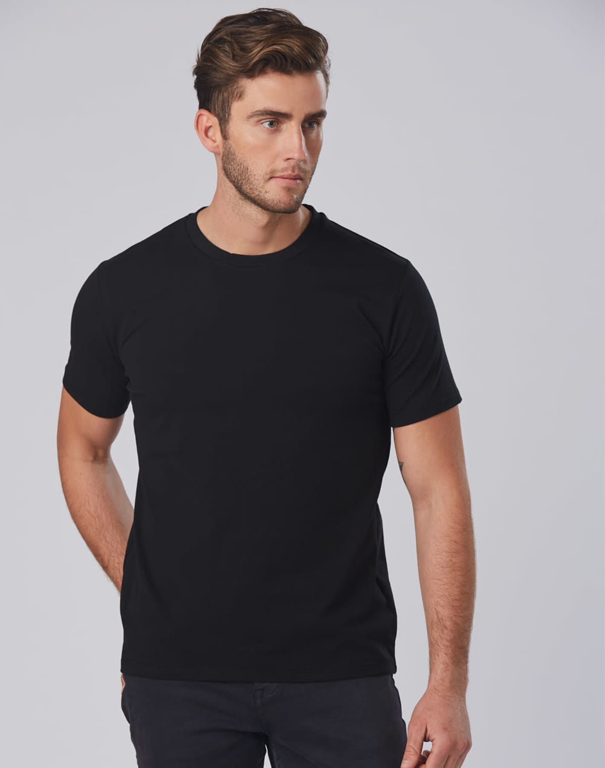 A man with short brown hair is wearing a plain black T-shirt and dark pants. He is standing against a plain grey background, gazing slightly to his right.
