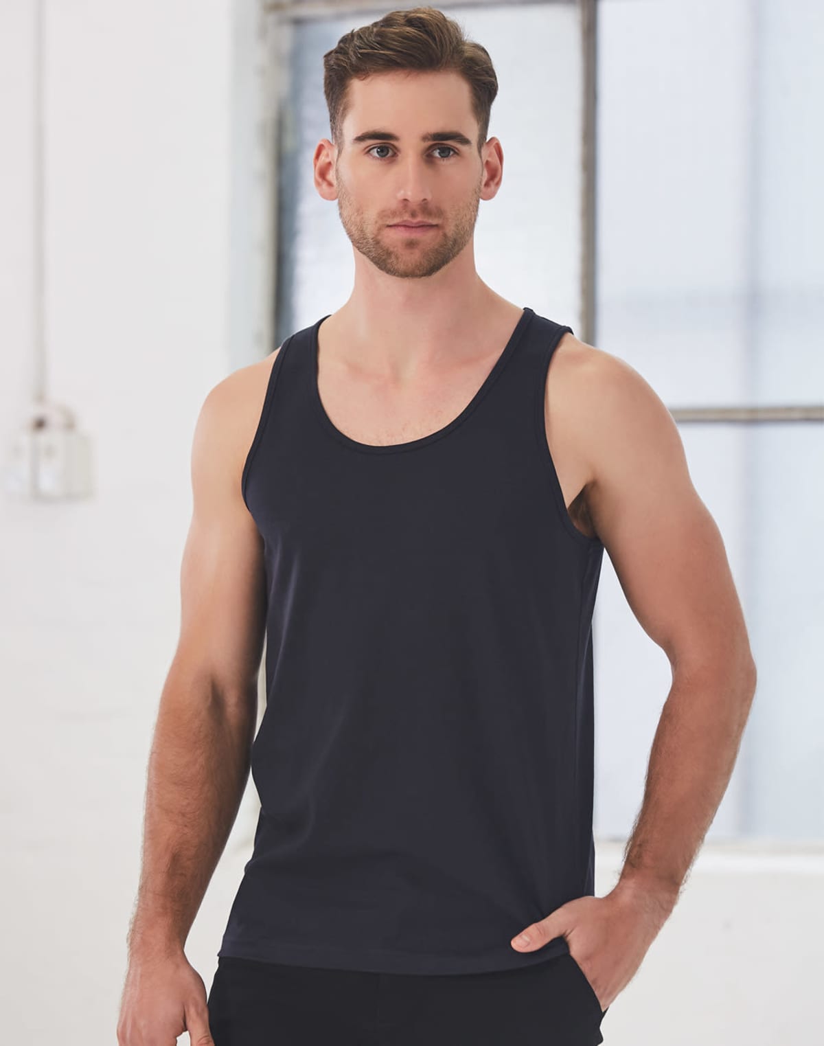 A man with short hair and a beard is wearing a black tank top. He stands in front of a large window with a neutral expression. The background is a light-colored, industrial-style wall.