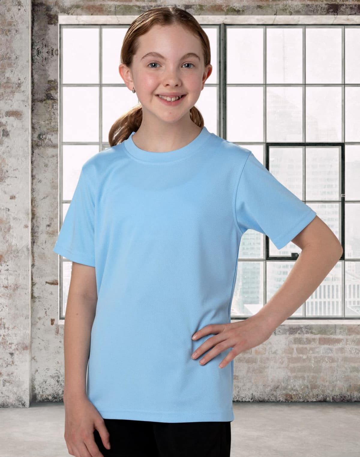 A young person with brown hair smiles while wearing a light blue T-shirt and black pants. They stand in front of a large window with a brick wall, letting in natural light.