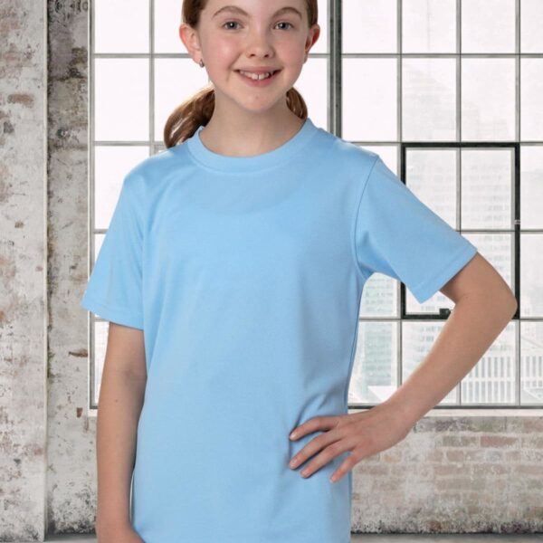 A smiling girl with brown hair is wearing a light blue T-shirt and black pants. She stands in front of a large window with a brick wall frame. The window shows a blurred cityscape in the background.